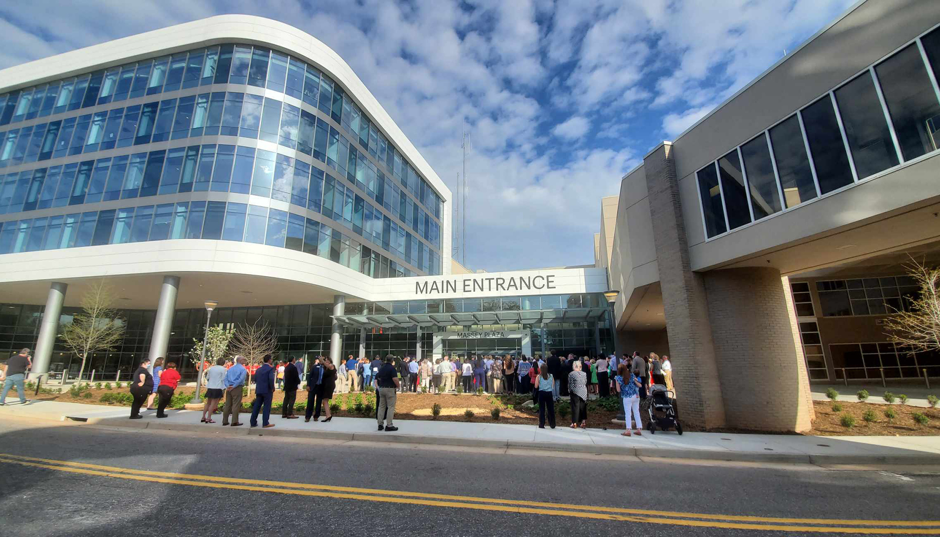 Ribbon cutting ceremony in front of new building. A good number of folks are watching the event. 
