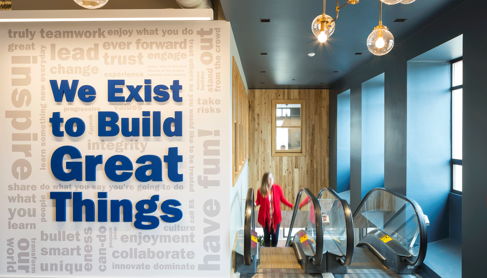 A woman walks up an escalator next to a wall that says "We Exist To Build Great Things"